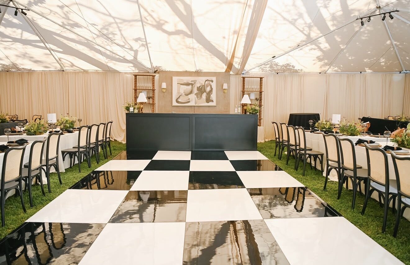 Black and white checkerboard dance floor under a draped tent, flanked by black cane dining chairs and a black bar