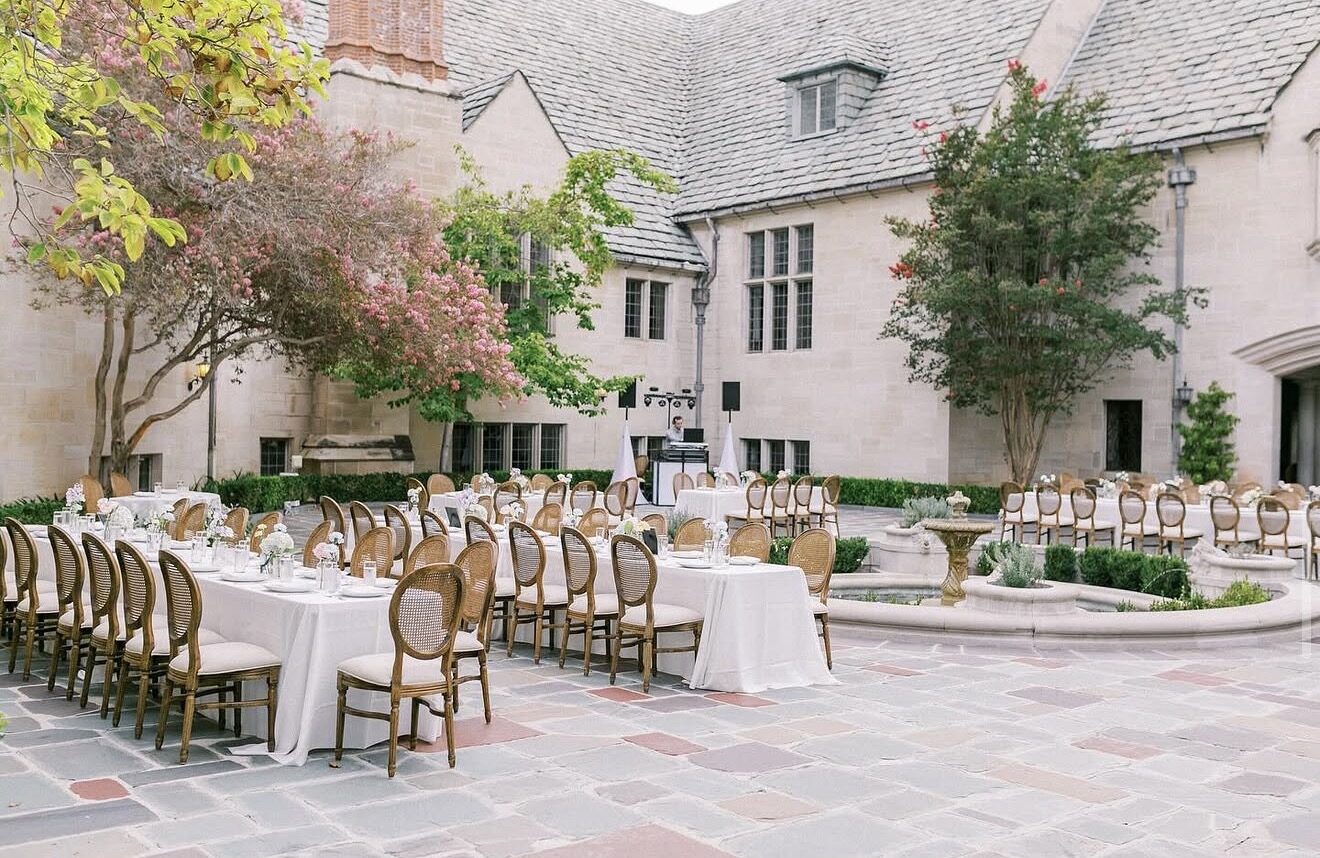 Long white dining tables and cane-back chairs in a stone mansion courtyard