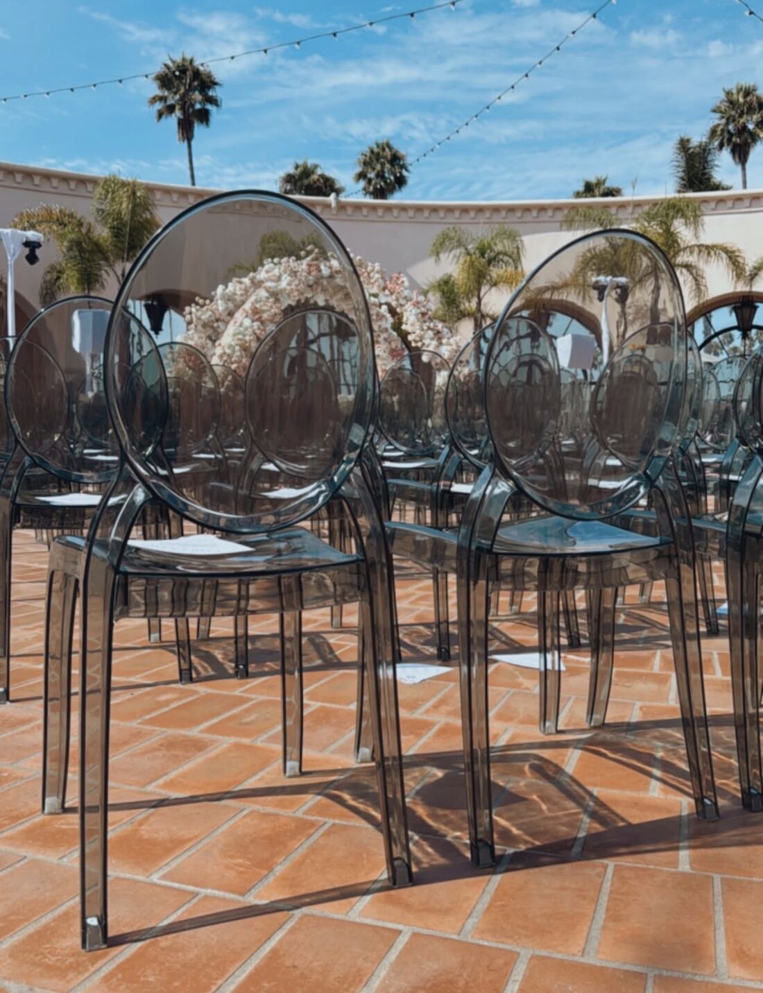 Rows of smoke acrylic ghost chairs on a terracotta courtyard before an outdoor ceremony