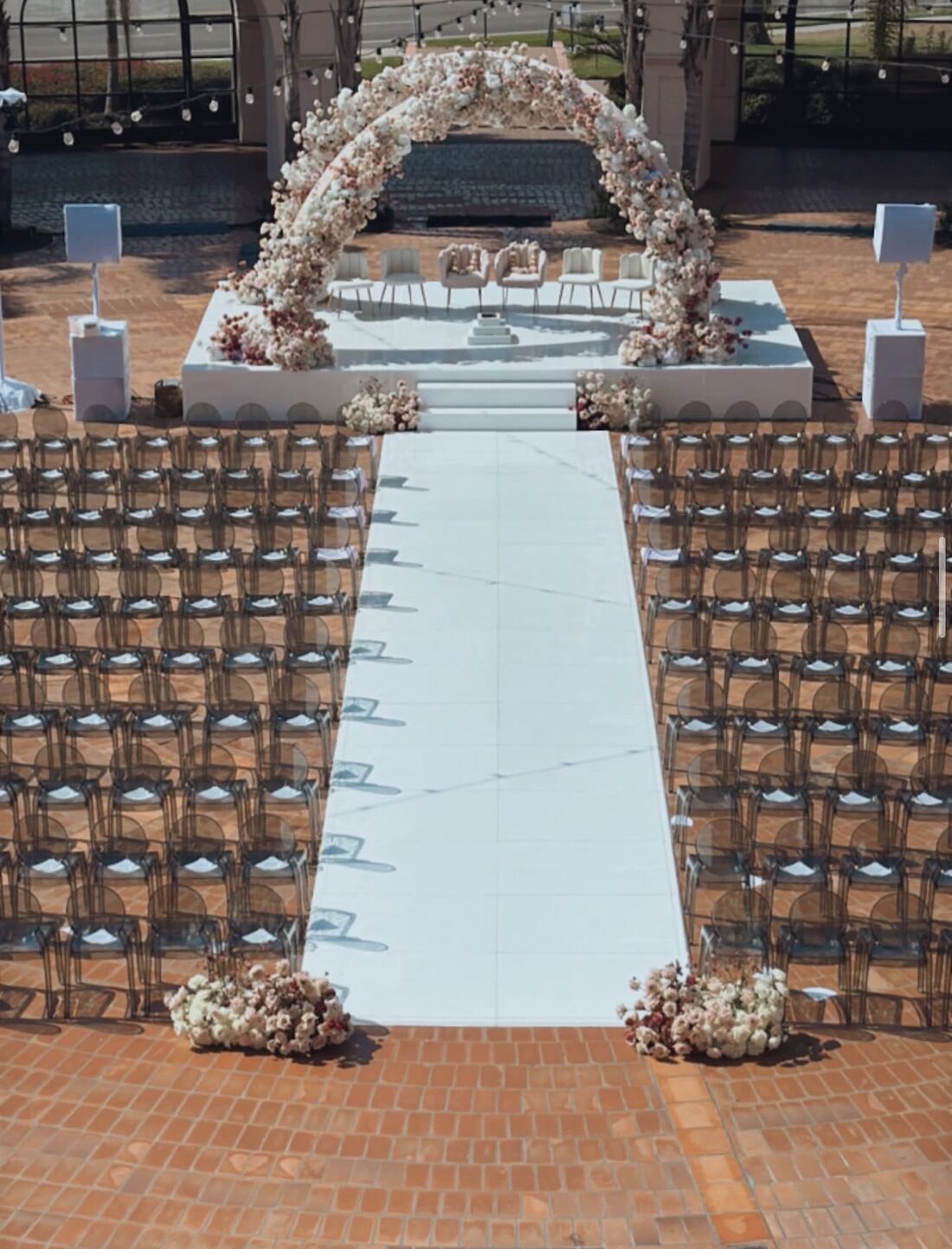 Aerial view of a white ceremony stage, floral arch and runway aisle lined with ghost chairs